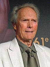 An older man is at the center of the image smiling and looking off to the right of the image. He is wearing a white jacket, and a tan shirt and tie. The number 61 can be seen behind him on a background wall.