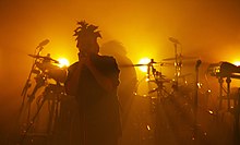 The picture depicts Tesfaye performing, with the lights giving the image an orange environment. In the background, there is a drum set.