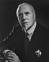 elderly white man with white receding hair and very small moustache and imperial beard, in contemporary lounge suit, facing the camera but not looking directly at it