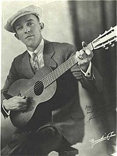 Autographed black and white picture of a man (Jimmie Rodgers) dressed in a striped suit, a white shirt, and wearing a tie and a beret. Sitting down, he holds the guitar to his chest resting the body on the right side and raising the neck while playing a C chord