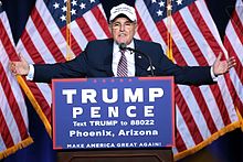 Giuliani with his arms outstretched, standing a podium with a banner for the Trump/Pence ticket