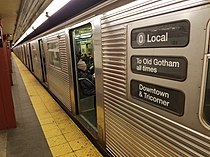 A corrugated silver metal subway train sits with its doors open in a station. Its rollsign reads "0 Local / To Old Gotham all times / Downtown & Tricorner".
