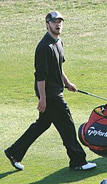 A young man dressed in black shirt and pants is walking while holding a golf stick