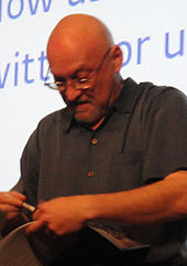 A caucasian, bald man wearing glasses and a dark shirt: He looks down at a book and pen held in his hands.