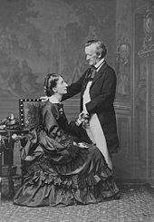 A couple is shown: On the left is a tall woman of about 30. She wears a voluminous dress and is sitting sideways in an upright chair, facing and looking up into the eyes of the man who is on the right. He is about 60, quite short, and balding at the temples. He is dressed in a suit with a tailcoat and wears a cravat. He faces and looks down at the woman. His hand rests on the back of the chair.