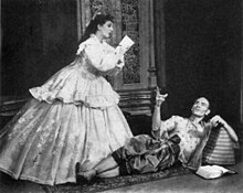 woman kneeling in front of a standing man; the two are conversing and each is gesturing with one hand as if ringing a small bell