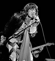 Mick Jagger sings onstage while holding a microphone stand up off the ground using both hands (left holding the pole and right the microphone body).