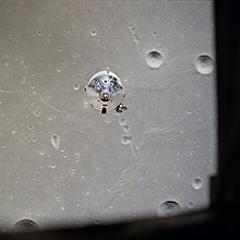 The top of the silvery command module is seen over a gray, cratered lunar surface