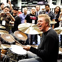 JR drumming at a convention, laughing as a crowd of musicians watches
