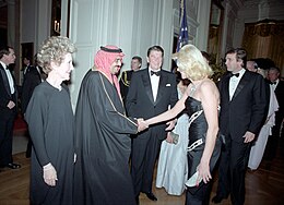 Ivana Trump and King Fahd shake hands, with Ronald Reagan standing next to them smiling. All are in black formal attire.