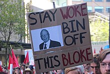 Cardboard sign at a street demonstration reading "Stay Woke – Bin Off this Bloke" with a picture of Rupert Murdoch