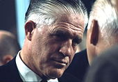 Headshot of a greying man in a suit who is indirectly facing the camera as he listens to a man a portion of whose head is shown from its side-to-rear