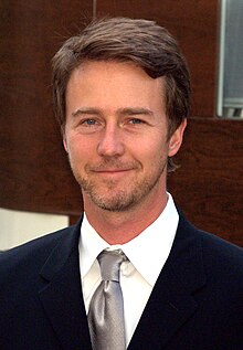 A portrait of Edward Norton, a blonde Caucasian man in a white plaid shirt. He is smiling towards the camera.