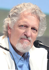 A white man with a short white beard, and white and grey curly hair: He wears a white suit with a blue shirt. The man stares away from the camera looking angry.