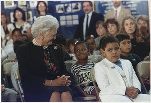 Barbara Bush and a young girl, seated next to each other in a room full of people and smiling at each other