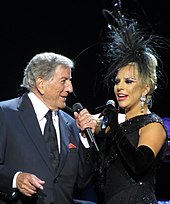 A man and a woman standing closely together. The man (left) is wearing a gray suit, white shirt and a black tie while the woman (right) is wearing a black gown, black gloves and a black headpiece. They both hold a microphone in their left hand.