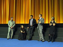 Scott Franklin, Mila Kunis, Vincent Cassel, Darren Aronofsky, and Sandra Hebron stand on a stage with a golden curtain backdrop wearing formal attire and discussing Black Swan