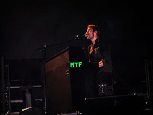 A short-haired man wearing a black outfit performs with a microphone and a piano