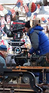 A man wrapped in a blue jacket and black hat looks down the scope of a large film camera. There is red, white, and blue bunting hanging overhead.