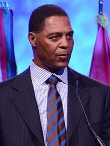 Marcus Allen in a suit jacket, dress shirt, and tie, speaking at a podium.