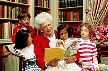 Bush sits in a chair and reads with four children surrounding her