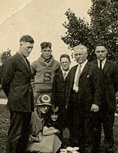 Five males of varying ages stand in a tight group, outdoors. Two sitting females huddle with them: a woman in a dress and a 1920s-style bonnet and a young girl in a dress. All have somber expressions. All the males wear jackets and suit ties with the exception of a teenage youth in a collared shirt and loop-collared, pullover sweater with a large block letter sewn onto the sweater's front.