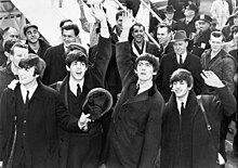 Black and white picture of the Beatles waving in front of a crowd with an set of aeroplane steps in the background