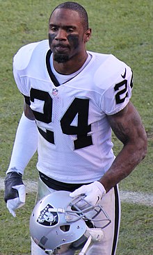 Charles Woodson wearing his Raiders uniform on a football field while carrying his helmet.