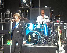 Two men on a stage. One is next to a microphone. The other is sitting behind a drum set.