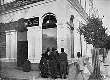 Photo of large white building with one sign saying "Moritz Schiller" and another in Arabic; in front is a cluster of people looking at a poster on the wall.