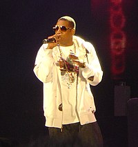 An image of an African American male wearing sunglasses with black pants and a white shirt and jacket. He is speaking into a microphone while holding it with his right hand. In the mostly dark background, a red colored wall with a design can be seen.