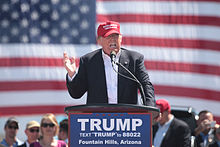 Trump speaking in front of an American flag behind a lectern, wearing a black suit and red hat. The lectern sports a blue "TRUMP" sign.