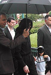 Jackson is wearing an overcoat and walking from left to right. His face is obscured by his hair. His son is wearing a mask and a baseball cap. Two men are with them; a third person is holding an umbrella over the Jacksons.