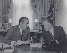 Two middle-aged men at a desk, in a forced pose of engaged conversation