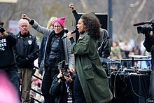 Alicia Keys photographed at the 2017 Women's March, dressed in winter clothing and raising her fist in the air alongside another woman on an outdoor stage with onlookers in the background