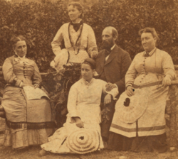 Portrait of five caucasian Latter-day Saints, married to each other in nineteenth-century Latter-day Saint polygamy, against the backdrop of what may be a hedge. All seem to be posing; none face the camera. Leftmost is a woman, seated, her hair done in a high, braided bun, wearing a dress with buttons down the middle; in her hands are an open book. Center-left, standing furthest to the back (though still very much with the portraited group) is a woman, her hair done up but resting low, in a polka-dotted top and a scarf or ascot around her neck and a skirt. She carries a hat, held to her waist. Center is a woman, sort of kneeling or seated (perhaps there is an unseen stool she's sitting on?). She wears a white dress, her hair is done up in a high and large bun and she wears a headband. In her right arm she holds a hat, over her knees; her left arm rests on the lap of the man sitting center right. She may be leaning against his legs. Center-right is a man, wearing a suit jacket of some kind and a high-collared shirt. He is balded and bearded. His left hand is placed over the left arm of the center woman. Rightmost is a woman, her hair done up but resting low, sitting in a visibly wooden (likely handcrafted) chair. She wears a dress with buttons going down the middle. She holds a hat, which looks very like center's hat, over her knees.