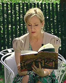 Frontale Farbfotografie einer Frau, die auf einem Stuhl sitzt und in ihren Händen ein offenes Buch hält. Sie hat ihre blonden Haare zusammengebunden und trägt eine kurzärmelige, graue Weste über einem blau-grünen Kleid. An ihrer linken Brust ist ein Ansteckmikrofon. Im Hintergrund ist ein Zaun mit Wiese.