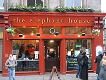 Frontale Farbfotografie von der roten Fassade des Cafés „the elephant house“. Flutlichter und Hängepflanzen zieren den Laden, der gut besucht ist. Rechts ist ein Hinweisschild über die Geburtsstunde der Harry Potter Bücher, das von Passanten teilweise verdeckt wird.