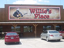 A store with a sign that reads "Willie's Place". The apostrophe is replaced in the sign by a bullet hole. The structure of the store is constructed in wooden with three columns. There are four windows and there are a red and a grey car in the parking lot.