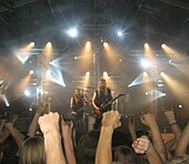 Image shows a band onstage with fans visible in the front of the picture. Some fans are raising their fists and others are raising their hands with the index finger and pinky extended.