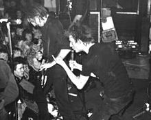 Two members of the rock band Crass are shown at a performance. From left to right are an electric guitarist and a singer. Both are dressed in all-black clothing. The singer is making a hand gesture.