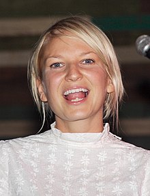A fair woman with short, bleached hair wears a modest white top as she looks into the camera, mouth open; a microphone is seen at the edge of the photo.