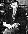 A photograph of Saul Bellow with an open book before a bookcase. He is wearing a suit and has somewhat curly hair.