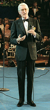 An elderly Stewart standing in a tuxedo on a stage, holding a microphone