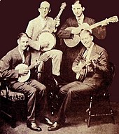 Black and white studio portrait of four men smiling while holding musical instruments: Three men play banjos and one of the back to the right plays a guitar. Jimmie Rodgers is seen second from the left wearing glasses and wearing a tan suit while holding up his banjo on his right knee as he raises his right leg on a chair. The two other men in the front sit in chairs facing each other sideways