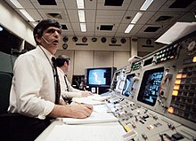 View along the computers banks in the mission control center and a flight controller sitting in front of a terminal