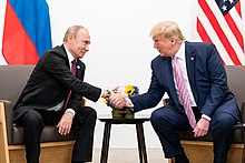 Trump and Putin, both seated, lean over and shake hands