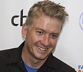 A Caucasian man stands in front of a poster background featuring different logos and smiles while looking ahead. He has blonde-gray hair and is wearing a blue crew-neck shirt underneath a black dress shirt