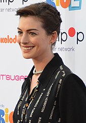 A short-haired brunette, wearing a black dress, is smiling to her left