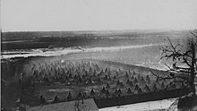 Black and white photo of one end of an island covered with hundreds of teepees inside a stockade